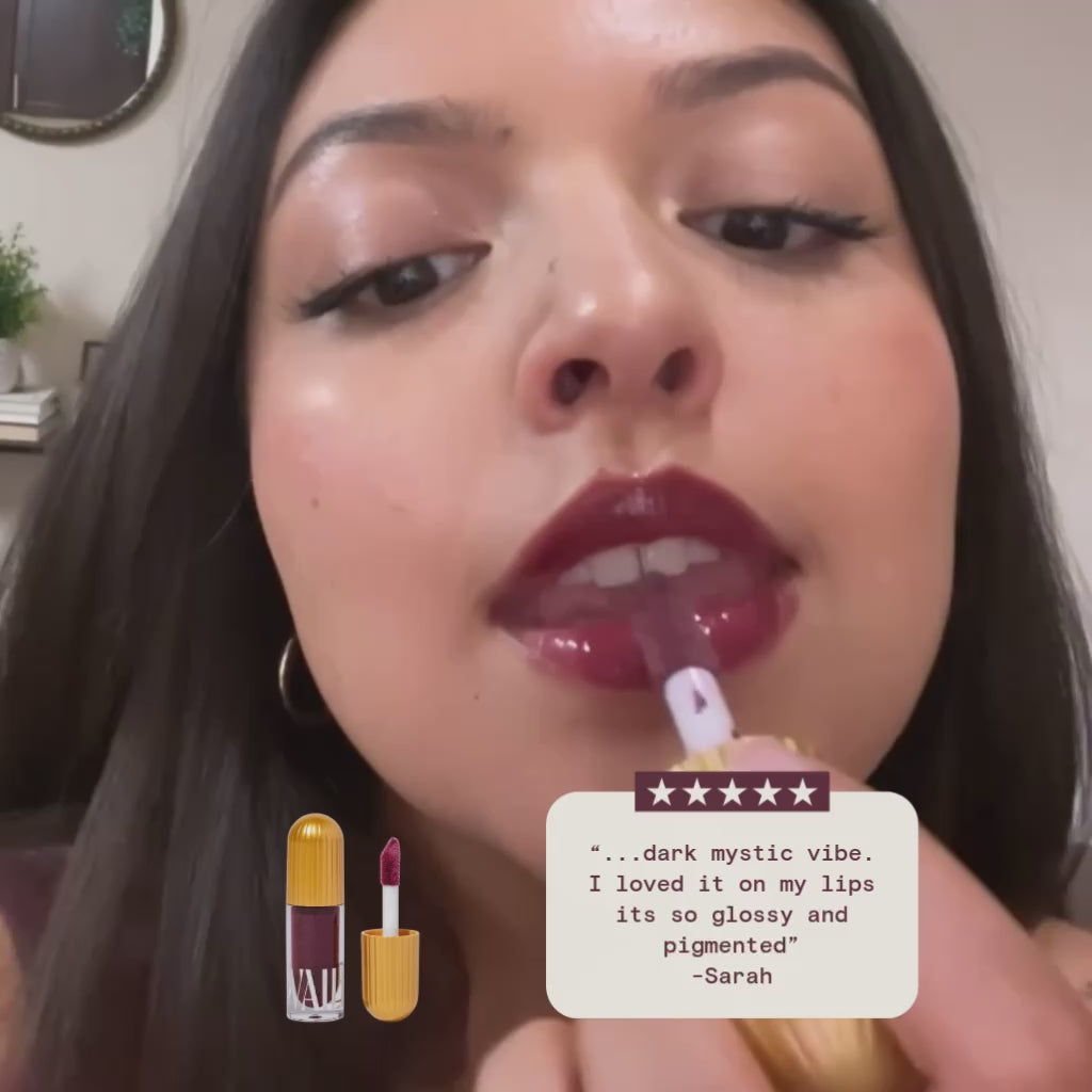 close up of a woman applying cleopatra lip gloss with a review on display that says "dark mystic vibe. I loved it on my lips its so glossy and pigmented" by sarah